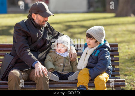 Vater sitzt auf der Bank im Park mit zwei Söhnen Stockfoto
