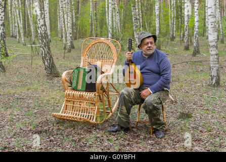 Senior Woman ist die Erholung im Birkenwald, sitzen auf einem Hocker Wicker und Mandoline halten Stockfoto