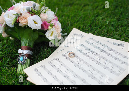 Zwei Trauringe auf einem Blatt Papier mit Noten. Brautstrauß auf dem grünen Rasen. Hochzeitskonzept. Stockfoto