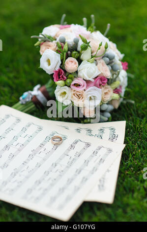 Zwei Trauringe auf einem Blatt Papier mit Noten. Brautstrauß auf dem grünen Rasen. Hochzeitskonzept. Stockfoto