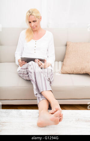 Eine reife Frau im Schlafanzug mit einem Tablet-PC auf dem Sofa. Stockfoto