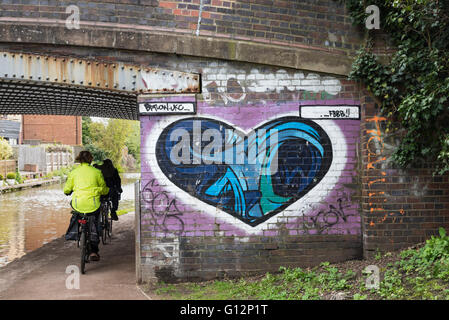 Frauen-Radfahrer, Radfahren auf dem Grand Union Canal Treidelpfad vorbei ein Herz geformt Graffiti-Kunst auf der Brücke Wand, England, UK. Stockfoto