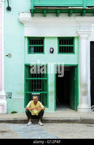 Ein Mann sitzt auf den Bordstein Rauchen einer Zigarette außerhalb eines Gebäudes mit einem Porträt von Che Guevara, Alt-Havanna, Havanna, Kuba Stockfoto