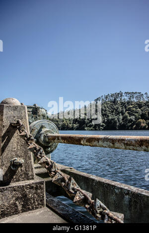 Verrostet, Maschinen und Kette von der Schwerkraft zugeführt Verdammung auf dem Fluß Mattupetty mit ruhigen See und Bäumen hinter Stockfoto