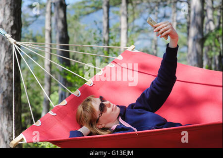 Lächelndes Mädchen in Gläsern in rote Hängematte im Wald genießen. Rothaarige Frau mit Sommersprossen lächelt, macht Selfie mit Smartphone. Stockfoto