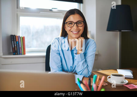 Lächelt freundlich attraktive Unternehmerin mit Brille sitzt an ihrem Schreibtisch in ihrem Büro zu Hause Stockfoto