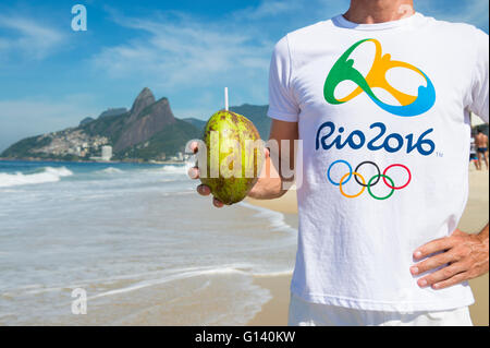 RIO DE JANEIRO - 10. März 2016: Mann trägt die offizielle Rio 2016 Souvenir steht T-shirt hält eine grüne Kokosnuss am Strand von Ipanema. Stockfoto