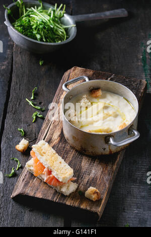 Vintage-Aluminium-Pfanne mit weiße Spargelcreme-Suppe mit Erbsen-Sprossen, gesalzener Lachs und Toast, serviert auf Schneidbrett aus Holz Stockfoto