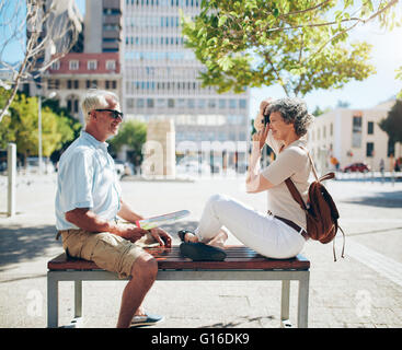 Seitenansicht der Reife Mann sitzt auf der Bank und Frau seine fotografieren mit Digitalkamera. Ältere Paare, die Spaß an ihrer vaca