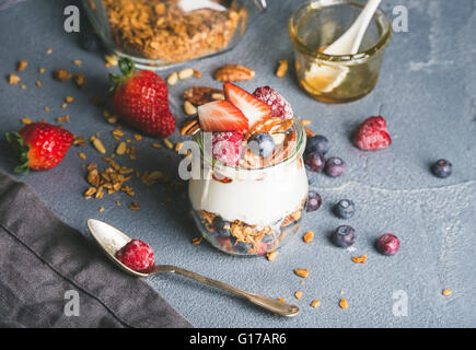 Joghurt-Hafer-Müsli mit frischen Beeren, Nüssen, Honig und Minze im Glas Glas auf grauem Beton strukturiertem Hintergrund, selektive fo Stockfoto