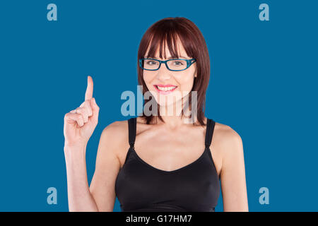Frau trägt ein Tank-Top und blaue Brillen mit dem Zeigefinger nach oben auf einem blauen Hintergrund Stockfoto