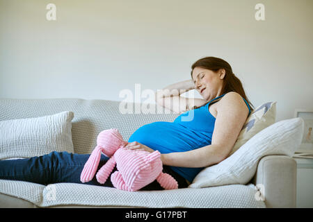 Seitenansicht der schwangeren Frau Holding rosa Plüschtier, geschlossenen Augen ruhen auf sofa Stockfoto
