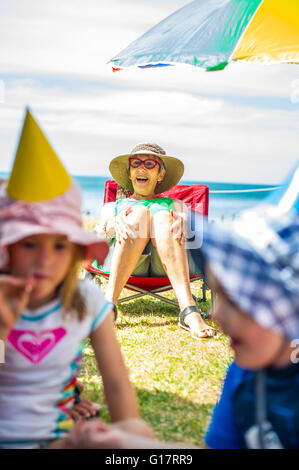 Kinder spielen bei Party vor senior Frau auf Liegestuhl am Küste, Waiheke Island, Neuseeland Stockfoto