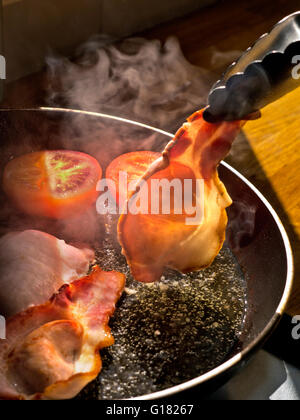 BACK BACON BRATEN Schaft von Sonnenlicht beleuchtet einen Raspel mit biologischem Bacon, der in einer heißen Pfanne mit heißem Ölrauch und Zange gedreht wird Stockfoto