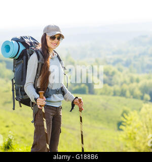 Junge lächelnde Frau mit Rucksack unterwegs grasbewachsenen Hügel. Fröhliches kaukasische Mädchen in Sonnenbrille genießen Sommerferien Stockfoto