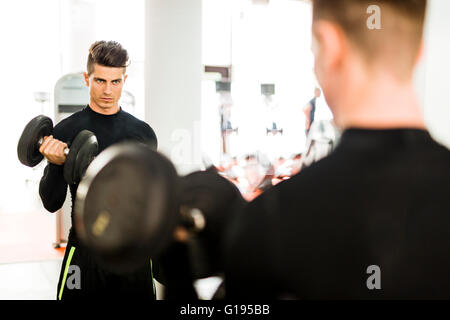 Muskulöser Jüngling, trainieren Sie im Fitnessraum und heben von Gewichten mit seiner Reflexion in einem Spiegel zeigen Stockfoto