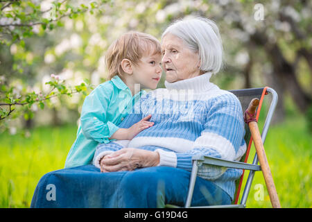 Kleiner Junge erzählt ein Geheimnis um seine Urgroßmutter in einen blühenden Obstgarten Stockfoto