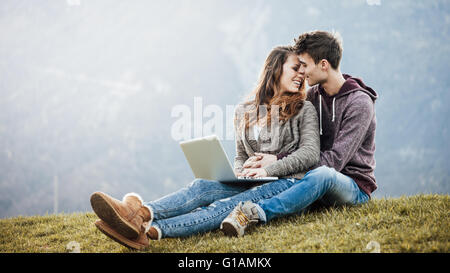 Junge romantisch zu zweit in der Natur auf dem Rasen sitzen, halten Sie einen Laptop und küssen Stockfoto
