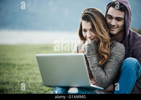 Verspielte junge Paar sitzen auf dem Rasen im Freien, umarmt und social Networking mit einem Laptop, Liebe und Beziehungen-Konzept Stockfoto