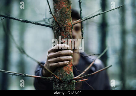 Junger Mann versteckt in den Wäldern und halten den Stamm, die hand auf Vordergrund, selektiven Fokus