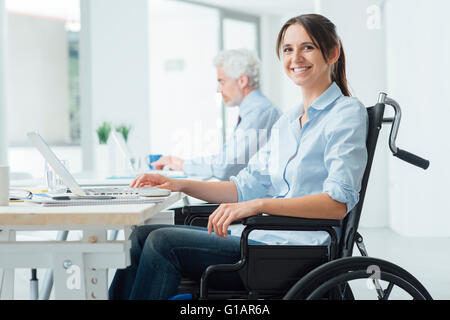 Sicher glücklich Geschäftsfrau im Rollstuhl am Schreibtisch arbeiten und einen Laptop verwenden, sie lächelt in die Kamera, Behinderung über Stockfoto