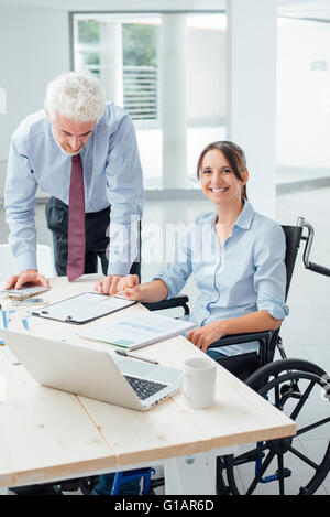 Zuversichtlich, Business-Frau im Rollstuhl mit ihr männlicher Kollege am Schreibtisch arbeiten und lächelt in die Kamera Stockfoto