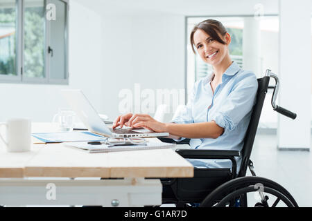 Sicher glücklich Geschäftsfrau im Rollstuhl am Schreibtisch arbeiten und einen Laptop verwenden, sie lächelt in die Kamera, Behinderung über Stockfoto