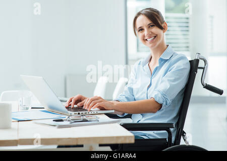 Sicher glücklich Geschäftsfrau im Rollstuhl am Schreibtisch arbeiten und einen Laptop verwenden, sie lächelt in die Kamera, Behinderung über Stockfoto