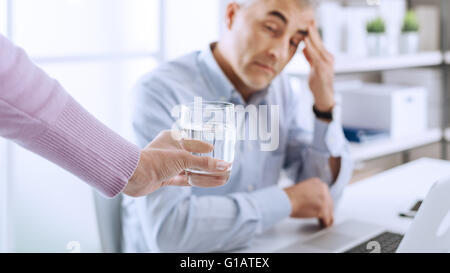 Müde Geschäftsmann im Büro am Schreibtisch arbeiten und haben eine starke Kopfschmerzen, sein Kollege ihm ein Glas Wasser und Touc gibt Stockfoto