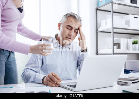 Müde Geschäftsmann im Büro am Schreibtisch arbeiten und haben eine starke Kopfschmerzen, sein Kollege ihm ein Glas Wasser und Touc gibt Stockfoto