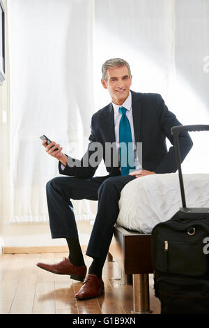 Business-Mann mit Koffer und Smartphone auf Bett in einem Hotelzimmer sitzen Stockfoto
