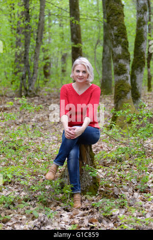 Schöne Blondine in einen roten Pullover sitzt auf einem Baumstumpf im Wald Stockfoto