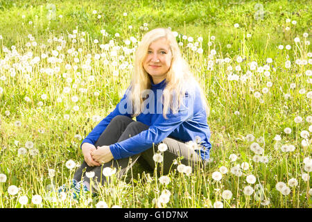 Blonde Frau mit langen Haaren sitzt auf Wiese mit weißen Löwenzahn Stockfoto