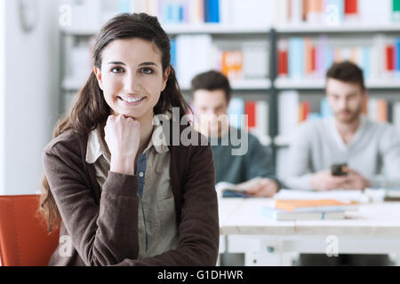 Jungen Student an der Bibliothek, Blick in die Kamera Lächeln, sitzen ihre Mitschüler am Schreibtisch Hintergrund auf Stockfoto