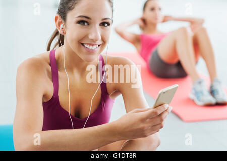 Junge lächelnde Frau in der Turnhalle eine Pause und anhören von Musik über ein smart Phone und Kopfhörer, Fitness und Jugend Konzeptionierung Stockfoto