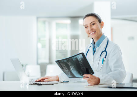 Lächeln zuversichtlich Ärztin am Schreibtisch sitzen und Untersuchung des Patienten Röntgen, sucht sie in die Kamera Stockfoto