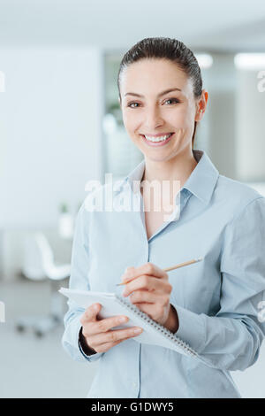 Junge professionelle Sekretärin aufschreiben von Notizen auf einem Notebook und lächelt in die Kamera, sie steht im Büro