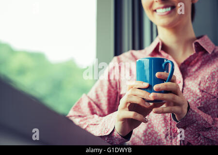 Frau, die einen erholsamen Kaffeepause am Fenster, sie lächelt und hält einen Becher Stockfoto