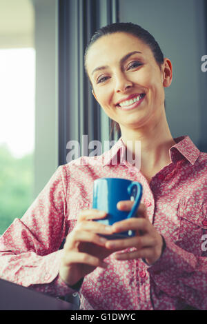 Frau, die einen erholsamen Kaffeepause am Fenster, sie lächelt und hält einen Becher Stockfoto