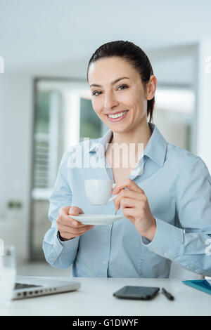 Lächelnde junge Frau hält eine Tasse Kaffee und eine Pause am Schreibtisch sitzen Stockfoto