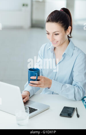 Lächelnde junge Frau hält eine Tasse Kaffee und eine Pause am Schreibtisch sitzen Stockfoto