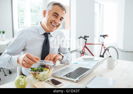 Glücklich Kaufmann brechen seinen Salat Pack öffnen und mit einem Mittagessen am Schreibtisch Stockfoto