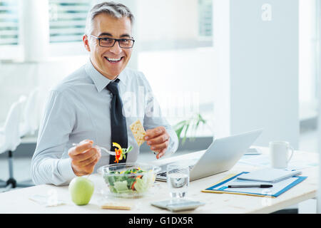 Lächelnd Geschäftsmann am Schreibtisch sitzen und mit einer Mittagspause, isst er eine Salatschüssel Stockfoto