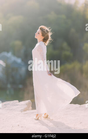 Schöne junge blonde Braut stehen in der Spitze des Hügels auf Natur mit fließenden weißen Kleid lächelnd Stockfoto