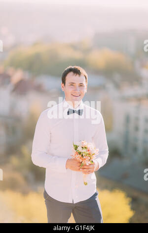 Stilvolle Brünette lächelnd Bräutigam in weißem Hemd mit blauen Bogen hält luxuriöse Blumenstrauß im freien Stockfoto