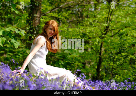 Eine schlanke, hellhäutige, junge Frau mit roten Haaren sitzt unter Bluebell Blumen im Wald Stockfoto