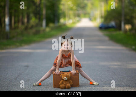 Lustiges kleines Mädchen mit einem Koffer und Teddybär auf der Straße. Stockfoto