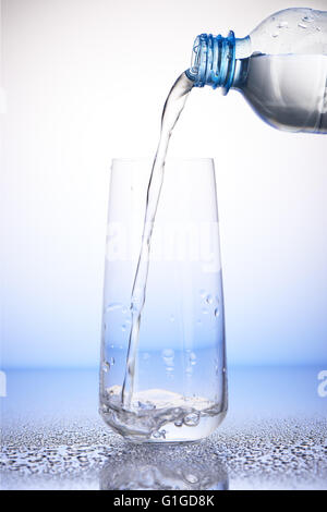 Wasser gießen aus Kunststoff-Flasche in ein Drittel voll Trinkglas mit Spiegelung auf Tropfen des Wassers auf weiß und blau Stockfoto