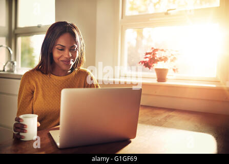 Erwachsenes Weibchen halten Tasse und lachen beim Betrachten ihrer Laptop-Computer beim Sitzen in der Nähe von hellen sonnigen Fenster in Küche Stockfoto