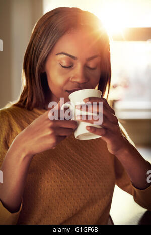 Attraktive junge schwarze Frau trinken eine Tasse frisch gebrühten Kaffee riechen den Duft mit ihren Augen geschlossen in Glückseligkeit als She-rel Stockfoto
